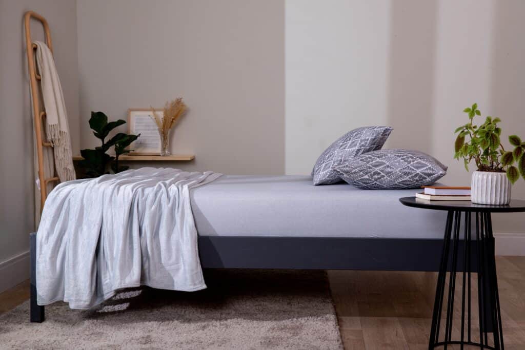 uma cama no centro de um quarto composta com um jogo de lençol cinza, na lateral há uma mesa de cabeceira com dois livros e uma planta, aos pés da cama uma mesa de estudos e uma escada com uma manta