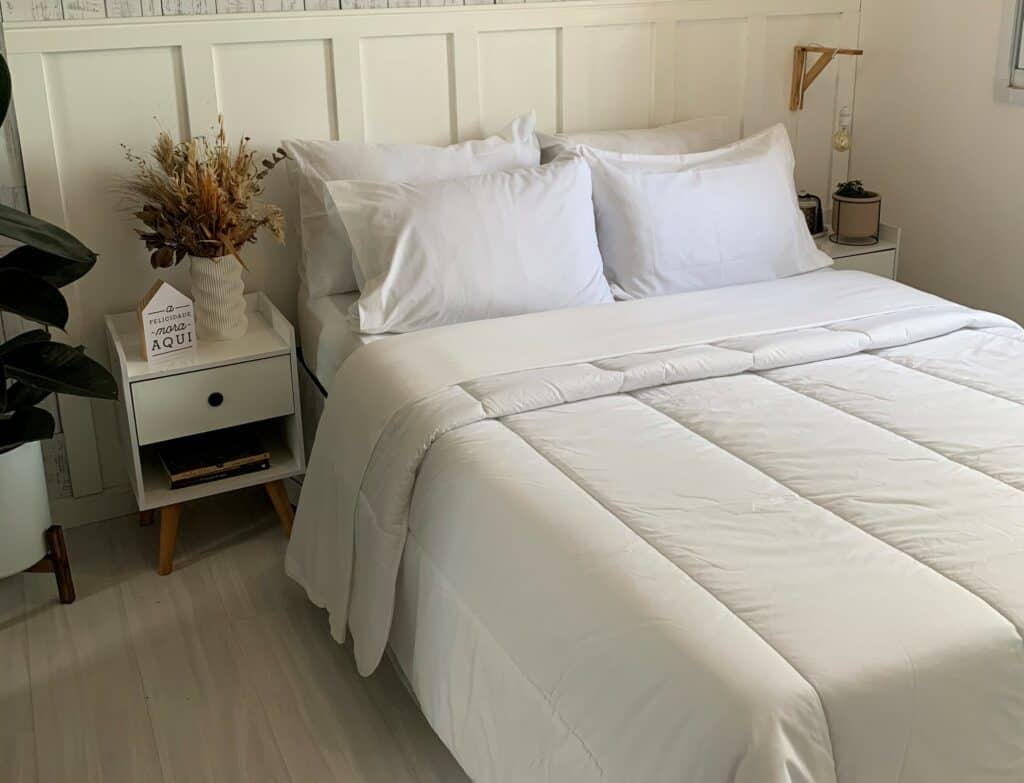 a decoração de quarto pequeno dessa imagem conta com roupas de cama brancas e lisas, duas mesas de cabeceira laterais e uma planta, mostrando beleza na simplicidade