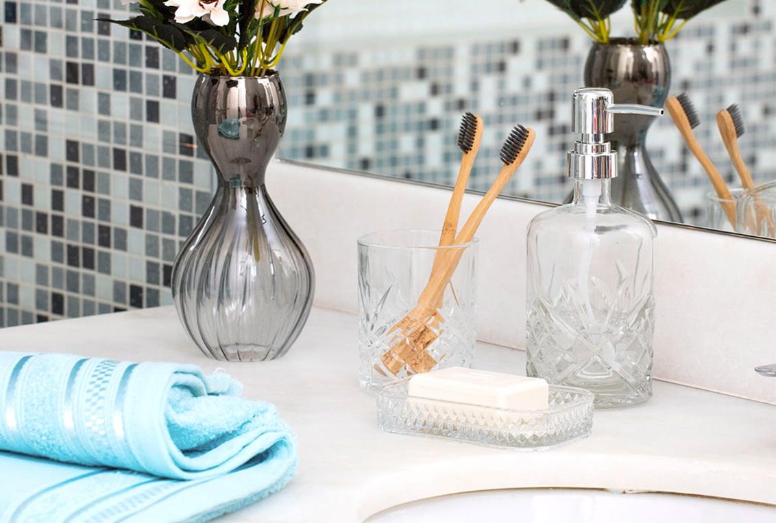 A imagem mostra uma bancada do banheiro, nela estão itens como kit lavabo com escovas e sabonete, toalha de lavabo, e um vaso com flores.