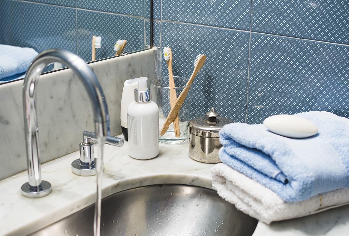 Na imagem centralizada há uma bancada com pia no banheiro, nela estão toalhas com um sabonete acima delas, copo com escovas de dentes e atrás deles está uma parede com azulejos decorados.