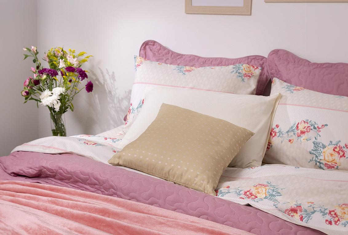Na imagem está centralizada uma cama posta, com travesseiros, almofadas, lençol, colcha e manta aveludada. À sua esquerda está um vaso com flores de espécies diferentes.